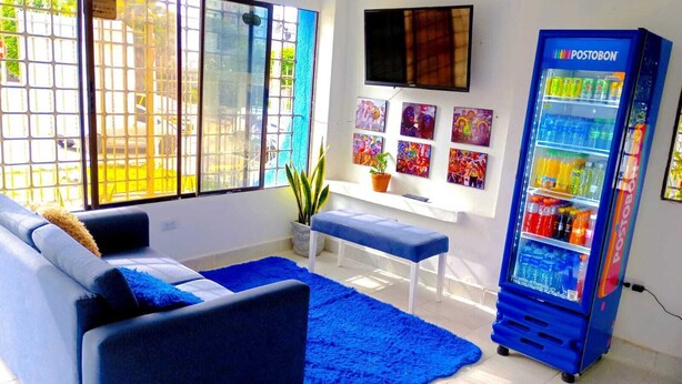 Een woonkamer met een bank, een lange hocker, een blauw vloerkleed, een televisie en een koelkast.