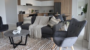 Ein modernes Wohnzimmer mit grauem Sofa, einem runden Couchtisch und einem grauen Sessel. Im Hintergrund befindet sich eine Küche mit Holzschränken und ein Essbereich mit Tisch und Stühlen.