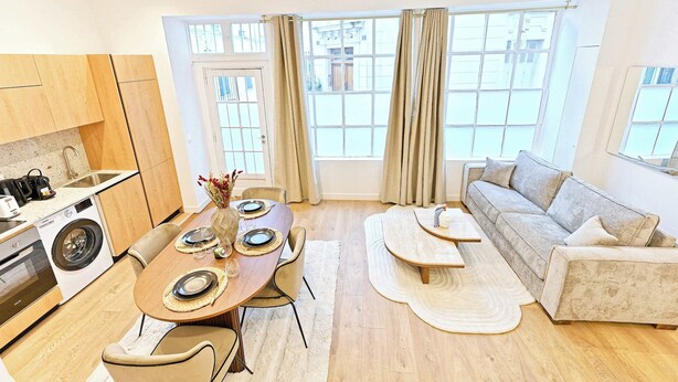 A modern kitchen with a built-in washing machine, a dining area with a round table and chairs, and a living area with a sofa and a large window. - Sublime air-conditioned flat Galeries Lafayette (Paris)