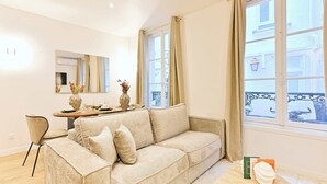 A modern living room with a beige sofa, a dining table with chairs, a vase with flowers, and a view of the street through large windows. - Magnificent air-conditioned flat Galeries Lafayette (Paris)