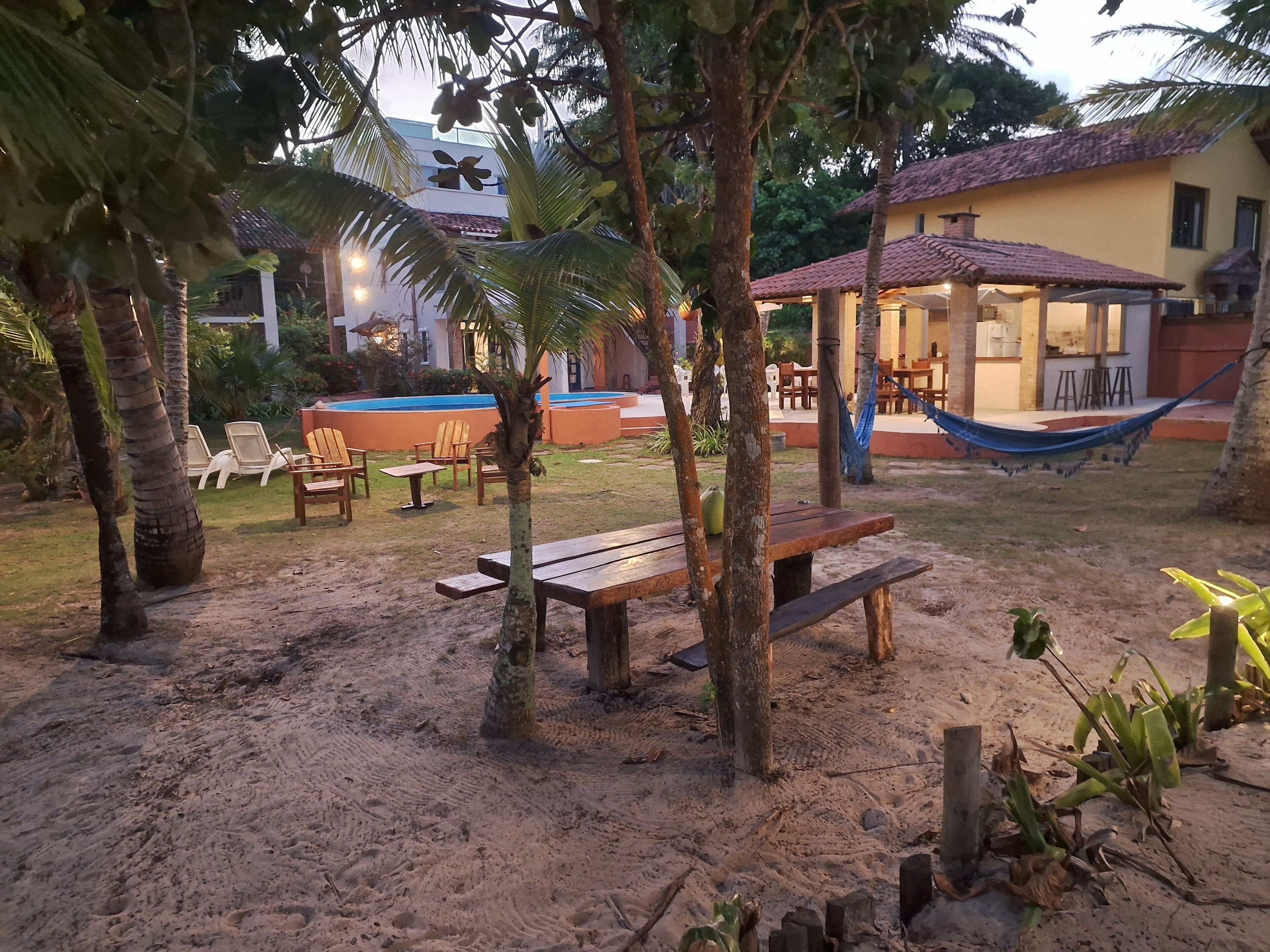 A sandy outdoor area with wooden benches, a pool, and a building with a tiled roof.