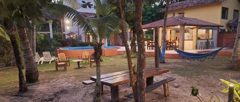A sandy outdoor area with wooden benches, a pool, and a building with a tiled roof.