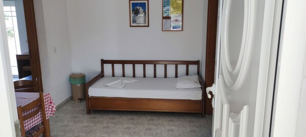 Une chambre à coucher avec un lit en bois, une petite table recouverte d’une nappe à carreaux, une poubelle verte et un tableau d’affichage accroché au mur.