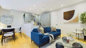 A modern living room with a blue sofa, a dining table with chairs, a kitchen area, and a large abstract painting on the wall. - Elegant Two-Bedroom Piccadilly Circus Apartment (Greater London)