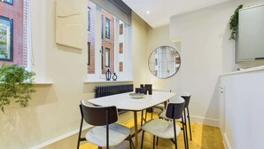 A modern dining area with a round table, four chairs, a large window with a view of a brick building, a radiator, and a mirror on the wall. - Elegant Two-Bedroom Piccadilly Circus Apartment (Greater London)