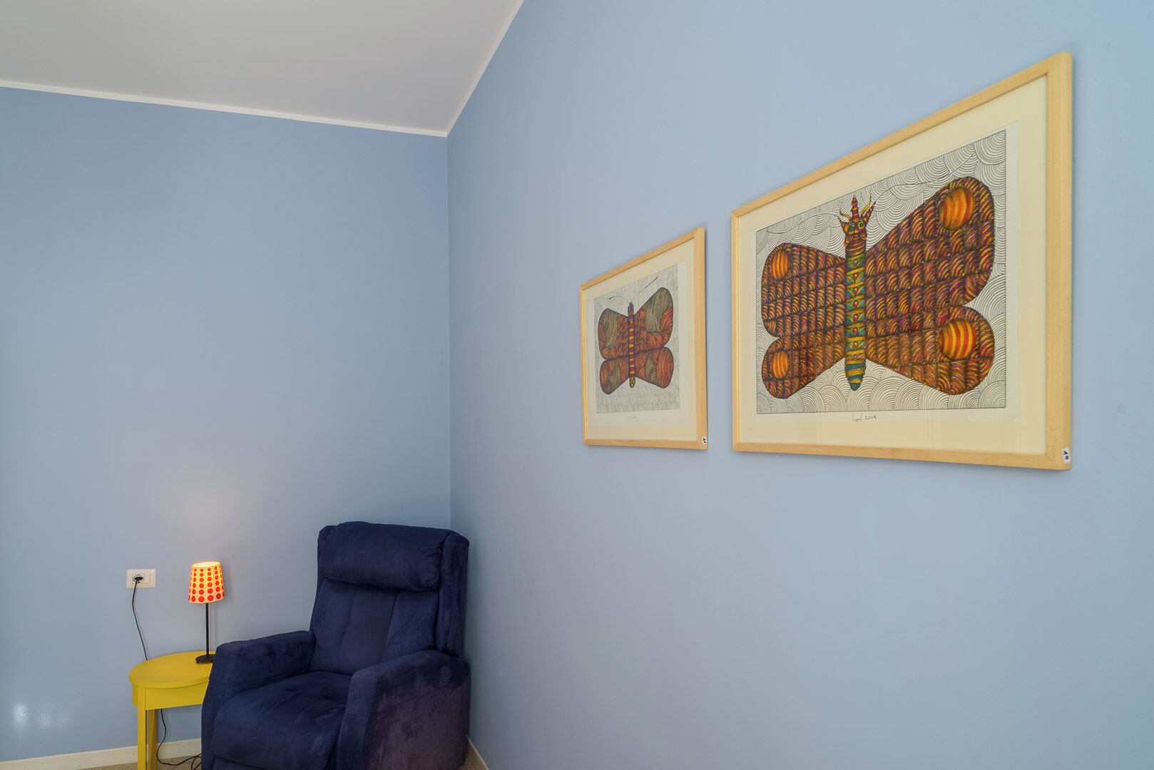 Une pièce aux murs bleu clair, avec un fauteuil bleu foncé et deux œuvres d’art encadrées représentant des papillons stylisés.