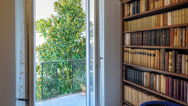 A room with a large bookshelf filled with books, a balcony with a metal railing, and a view of greenery outside.