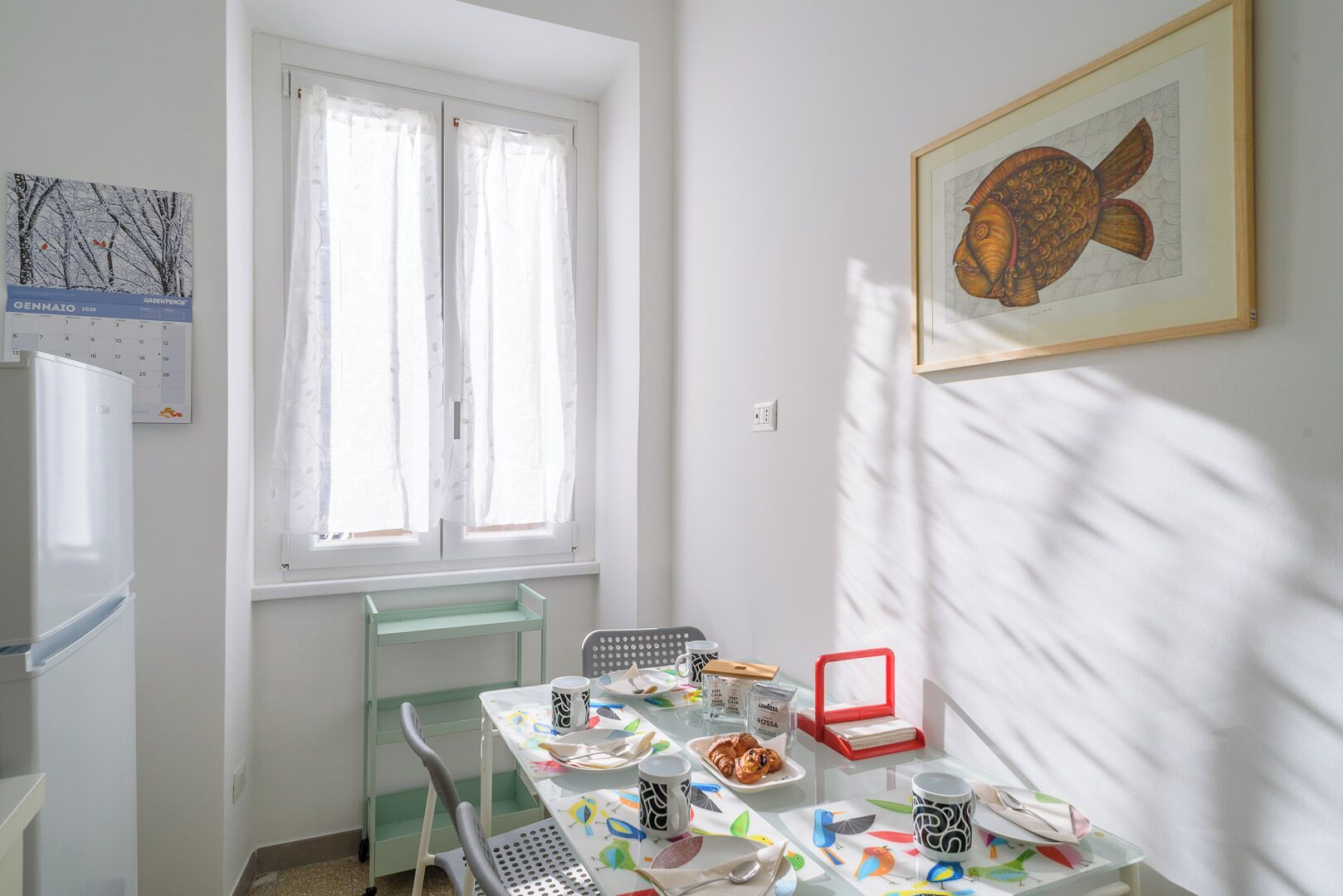A dining area with a table set for two, a window with sheer curtains, a framed artwork on the wall, and a refrigerator in the corner.