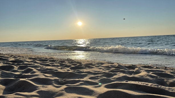 Una playa con arena y olas, cielo despejado y el sol poniéndose o saliendo.