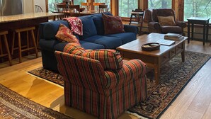 Ein geräumiges Wohnzimmer mit einem großen blauen Sofa, einem gestreiften Sessel und einem Holztisch. Mehrere Fenster sorgen für natürliches Licht und Blick auf den umliegenden Wald.