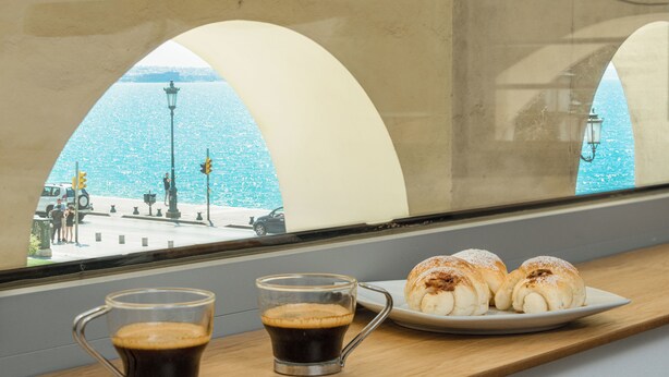A table with two glasses of coffee and a plate of pastries, viewed through a window with a view of the sea. - SQUARE Elegant Suite II on Aristotelous Square (Thessaloniki)