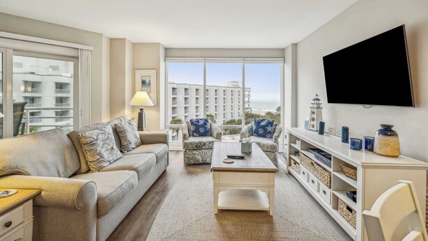 A living room with a sofa, armchair, coffee table, TV, and a view of buildings through large windows. - 2 BDR w/Water Views, Beach Access, & Shared Pool! (Hilton Head Island)