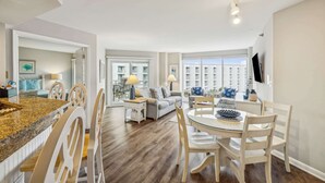 A modern living room with a dining area, large windows, and a flat-screen TV mounted on the wall. - 2 BDR w/Water Views, Beach Access, & Shared Pool! (Hilton Head Island)
