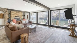 A spacious living room with a stone fireplace, leather sofa, and large windows overlooking a lake. - Foggy River Retreat – For 16! (Hollister)