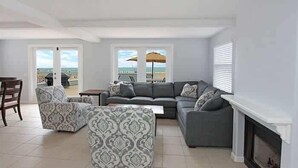 Ein Wohnzimmer mit grauem Ecksofa, einem gemusterten Hocker und einem Kamin. Ein Balkon bietet Blick auf den Strand.