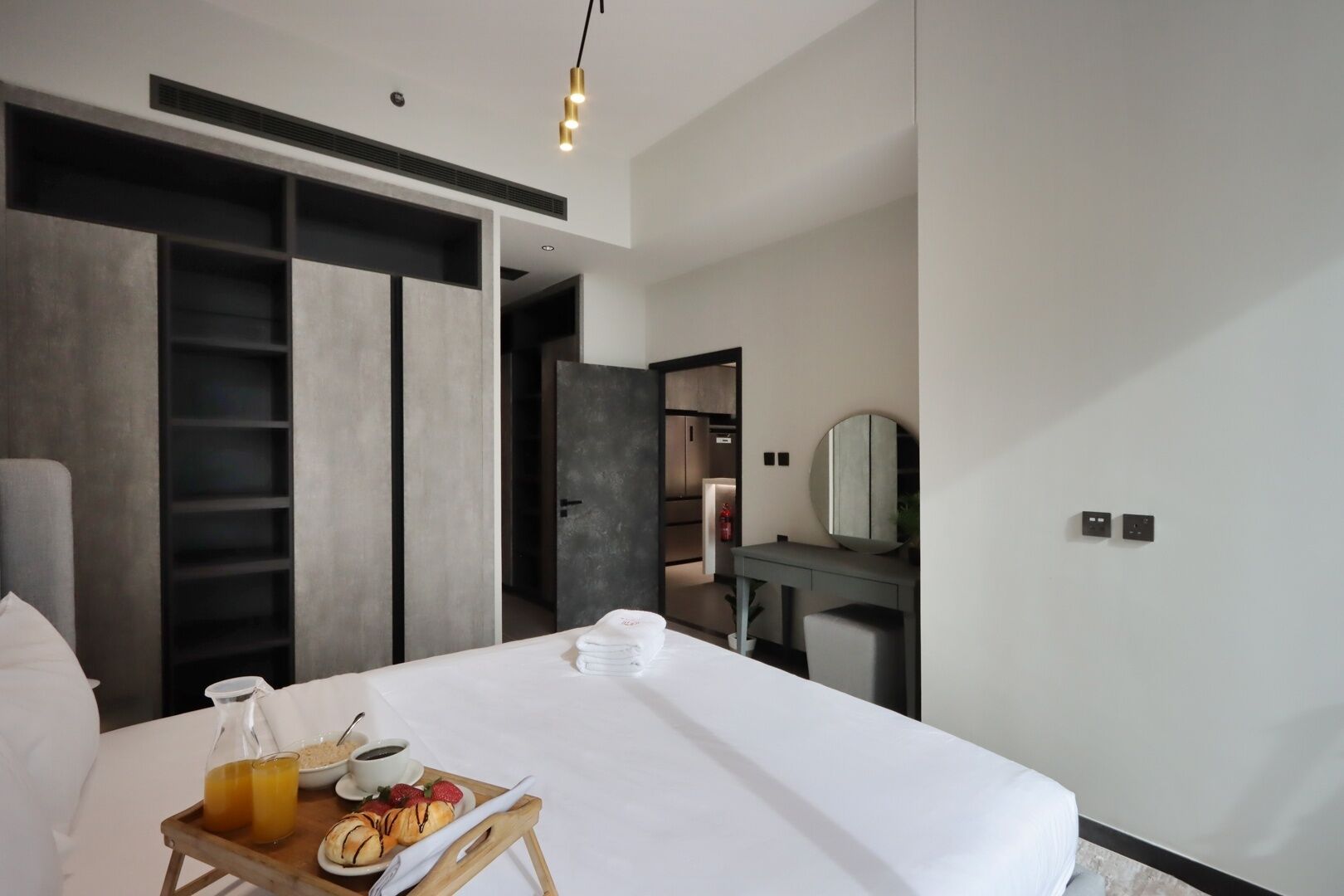 Ein modernes Hotelzimmer mit einem großen Bett, einem Nachttisch mit Frühstücksangeboten, einem Kleiderschrank und einem durch eine offene Tür einsehbaren Badezimmer.