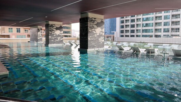Una piscina interior cristalina con pilares de mármol, sillones de descanso y vistas de edificios circundantes.