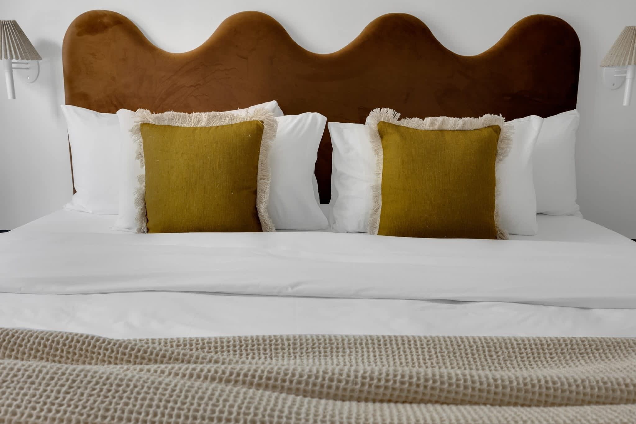 Ein Bett mit wellenförmigem Kopfteil, Kissen in Weiß und Senfgelb sowie einer gemusterten Decke.