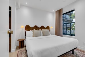 A neatly made bed with a wooden headboard and two bedside lamps, a window with curtains, and a small wooden stool beside the bed.