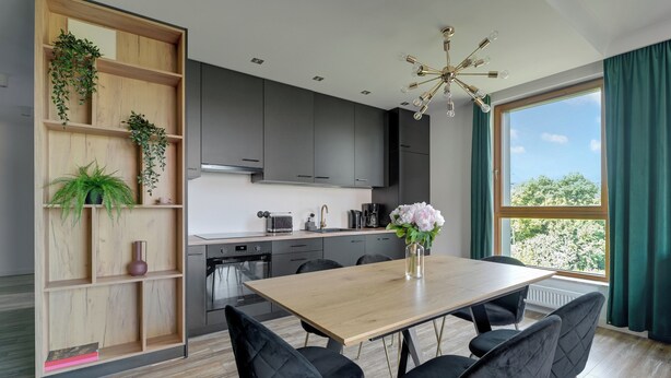 A modern kitchen with dark cabinets, a wooden dining table, and black chairs. There is a window with curtains, a ceiling light fixture, and a shelf with plants. - Lion Apartments - Front Park (Gdansk)