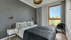A modern bedroom with a large bed, a nightstand, a chair, and a window with curtains. - Lion Apartments - Front Park (Gdansk)