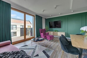 A modern living room with a large sliding glass door, a pink sofa, a round coffee table, a purple armchair, a wooden dining table, and a flat-screen TV mounted on the wall. - Lion Apartments - Front Park (Gdansk)
