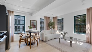 A modern living room with a white sofa, dining table, and chairs, large windows with curtains, and a kitchen area with a refrigerator. - Mercer-King Bedroom -Free parking (Montréal)