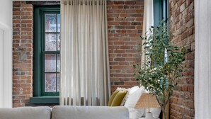 A cozy living room with a brick wall, a green-framed window, a beige sofa, a white lamp, and a potted plant. - Mercer- Studio 1Beds+Sofa bed / Free parking (Montréal)