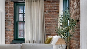 A cozy living room with a brick wall, a green-framed window, a beige sofa, a white lamp, and a potted plant. - Mercer- Studio 1Beds+Sofa bed / Free parking (Montréal)