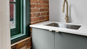 A kitchen with a brick wall, a window with a green frame, and a stainless steel sink with a gold faucet. - Mercer- Studio 1Beds+Sofa bed / Free parking (Montréal)