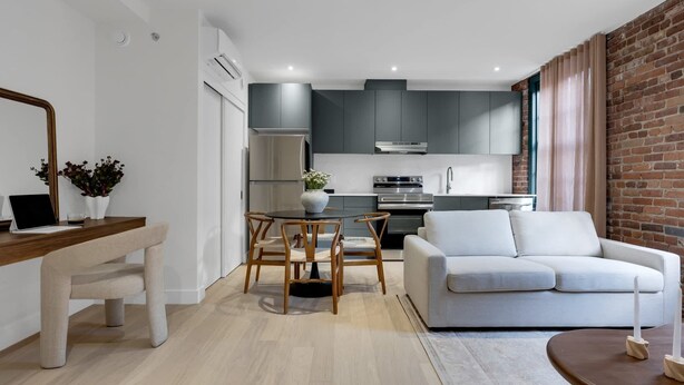 Una cocina moderna con gabinetes grises, un refrigerador de acero inoxidable y un horno. Hay un comedor con una mesa redonda y sillas, y un sofá blanco en la sala de estar.