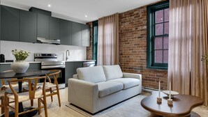 Una cocina moderna con armarios grises, una zona de comedor con una mesa redonda y sillas, un sofá beige y una pared de ladrillo.