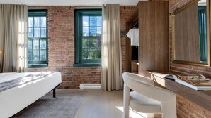 A modern hotel room with a brick wall, a large window with curtains, a bed, a desk with a mirror, and a chair.