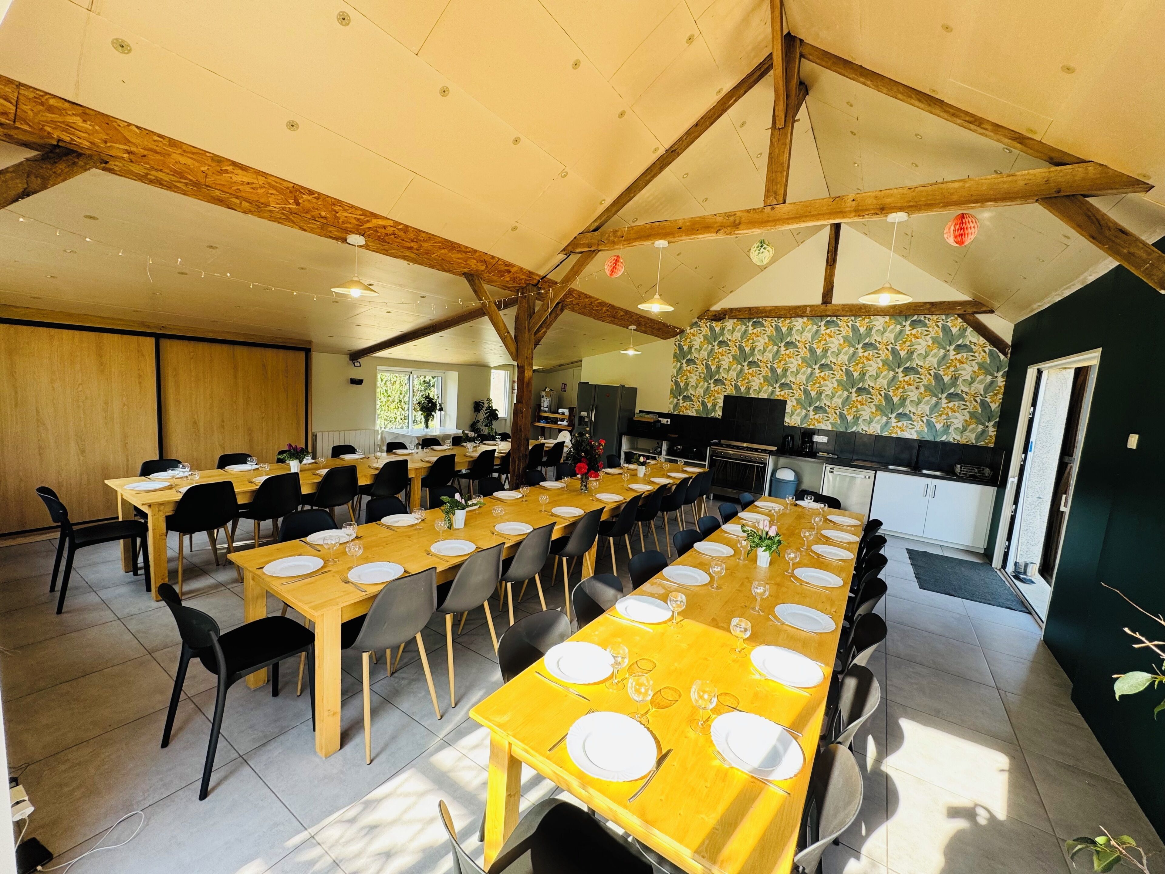 A dining area with multiple tables set for meals, a kitchen in the background, and a high ceiling with exposed beams.