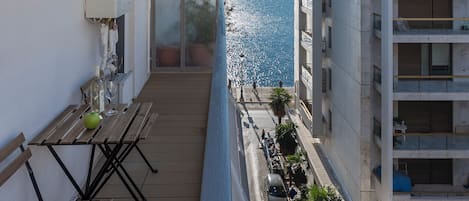 A balcony with a view of the sea, a wooden table with a green apple and a wine glass, and a building with balconies and a tiled facade.