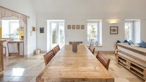 Un comedor amplio con una larga mesa de madera, sillas de madera y un banco con cojines. Hay cuadros en las paredes y vistas al exterior a través de grandes ventanales.