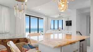 A modern kitchen with a marble countertop, white cabinetry, and a large window overlooking the ocean.