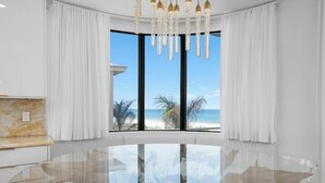 A spacious room with a large glass table, white curtains, and a view of the beach.