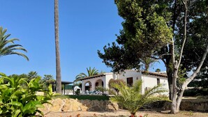 A white building with arches and a tiled roof, surrounded by palm trees and flowering plants. - LA CASITA | ideal villa for small families | 2 double bedrooms | private pool (Teulada)