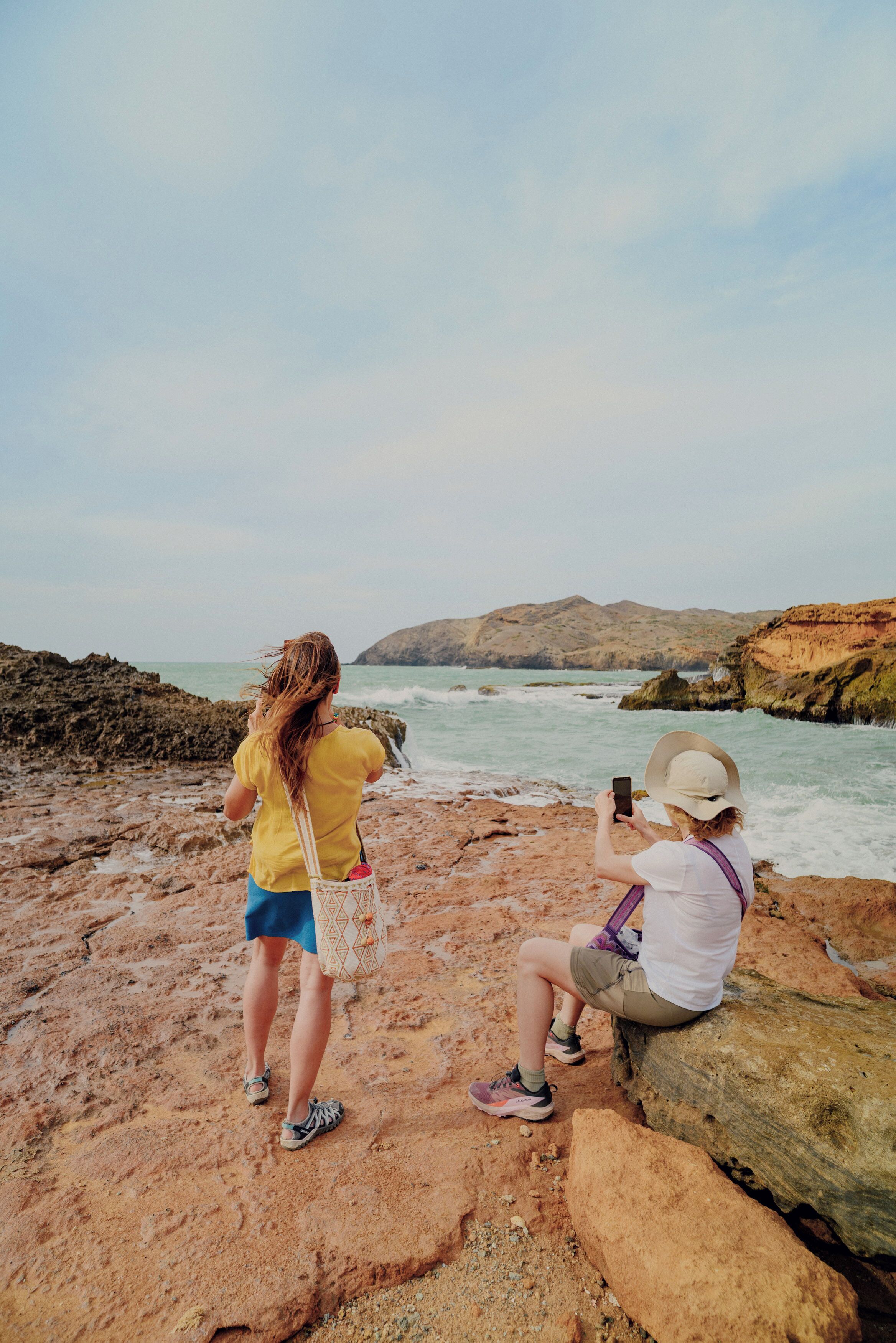 Two people on a rocky beach, one taking a photo, the other sitting and using a phone.