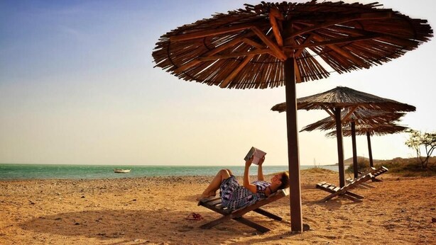 Een strandscène met iemand die op een ligbed ligt onder een rieten parasol, een boek lezend.