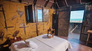 Een kamer met een bed, een klein tafeltje met een mand en uitzicht op het strand door een geopende deur.