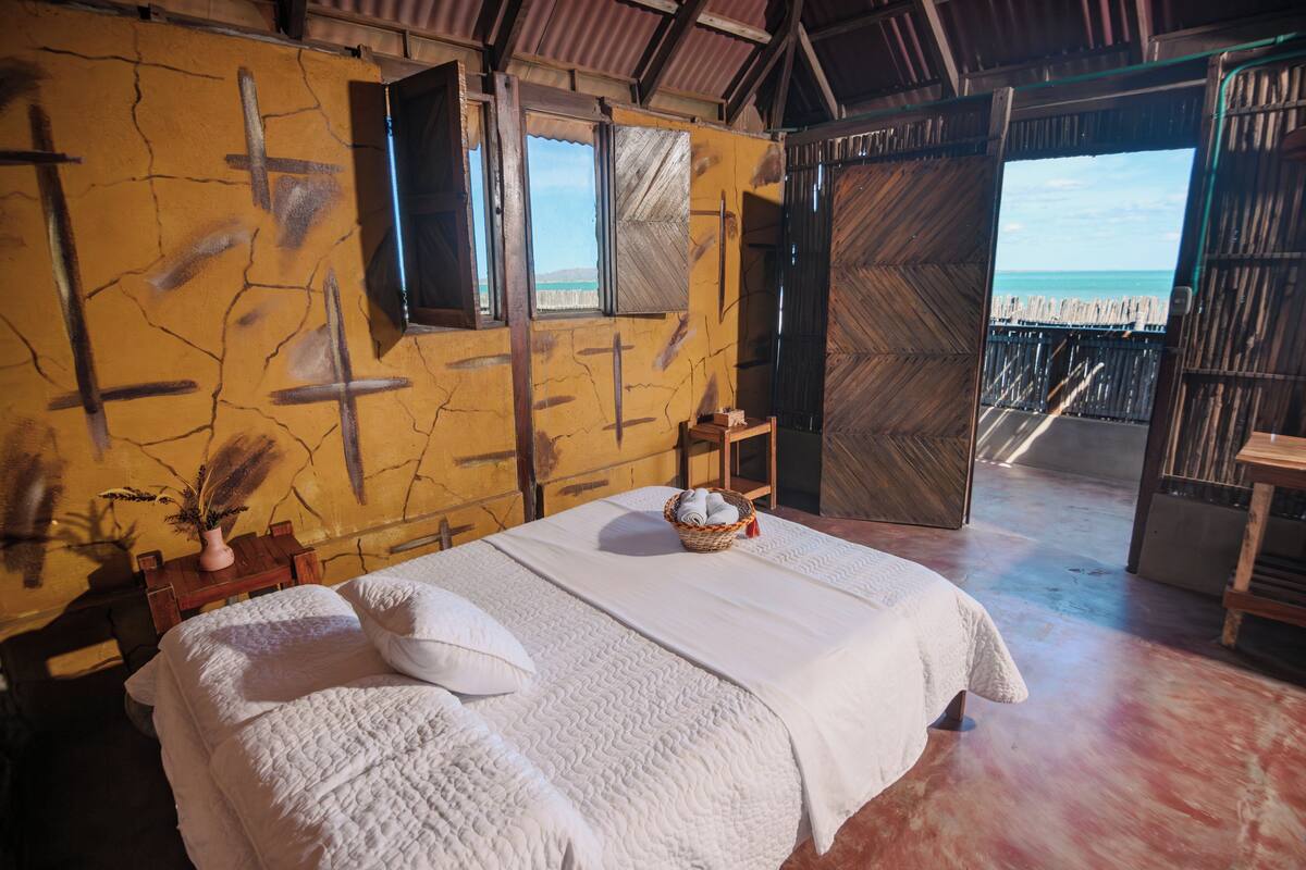 A room with a bed, a small table with a basket, and a view of the beach through an open door.