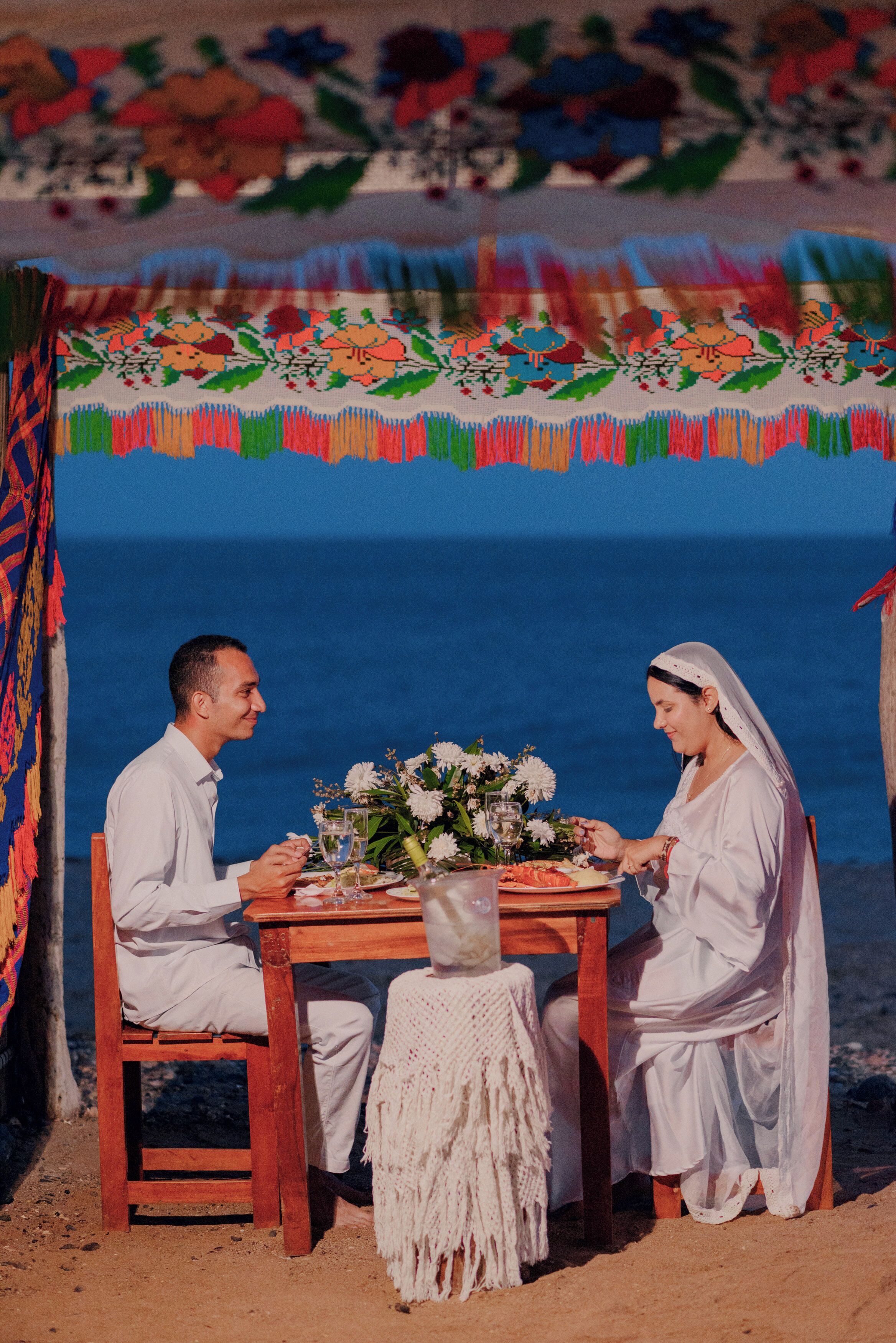 Una pareja cenando en una mesa de madera con un centro de mesa floral, frente a un fondo de playa y tela colorida.