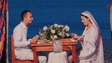 Una pareja cenando en una mesa de madera con un centro de mesa floral, frente a un fondo de playa y tela colorida.