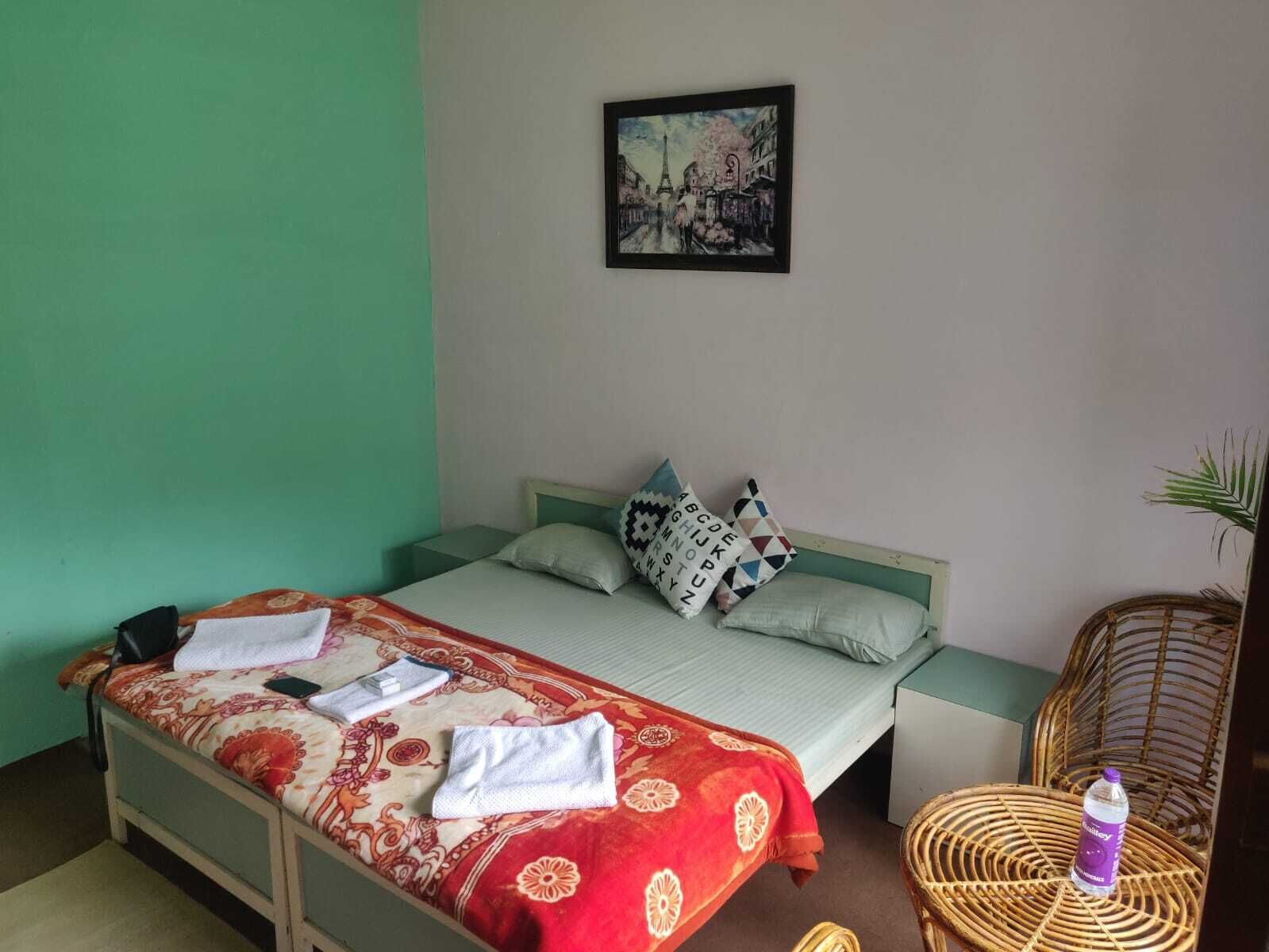 Quarto com cama, almofadas, um quadro na parede, uma cadeira de vime e uma garrafa de água.
