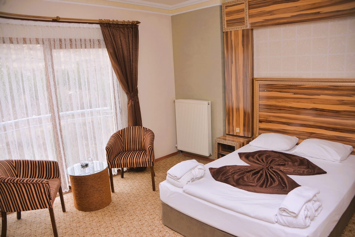 Une chambre d’hôtel avec un lit, deux fauteuils à rayures, une petite table ronde et une fenêtre ornée de rideaux légers.