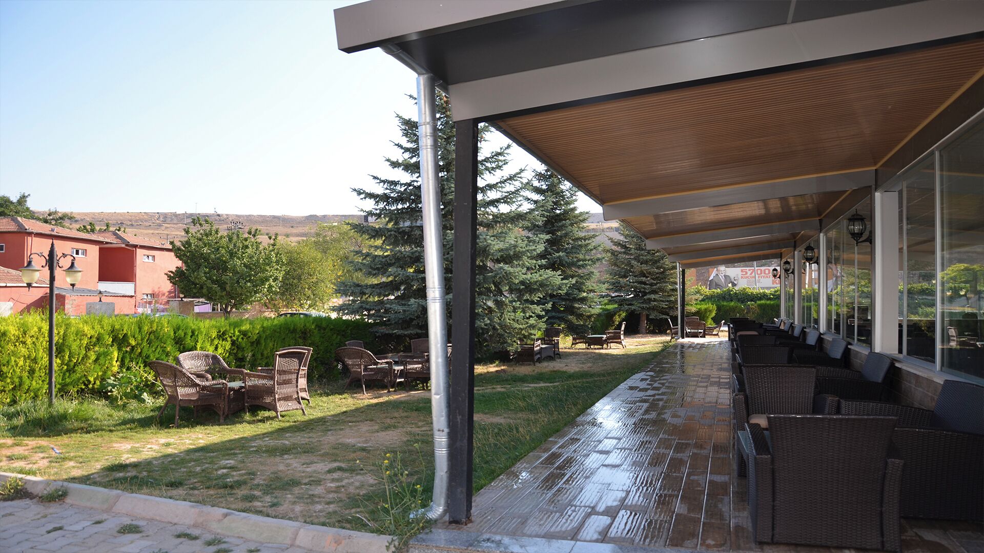 A covered outdoor seating area with wicker furniture, a wooden ceiling, and a view of a red building in the background.