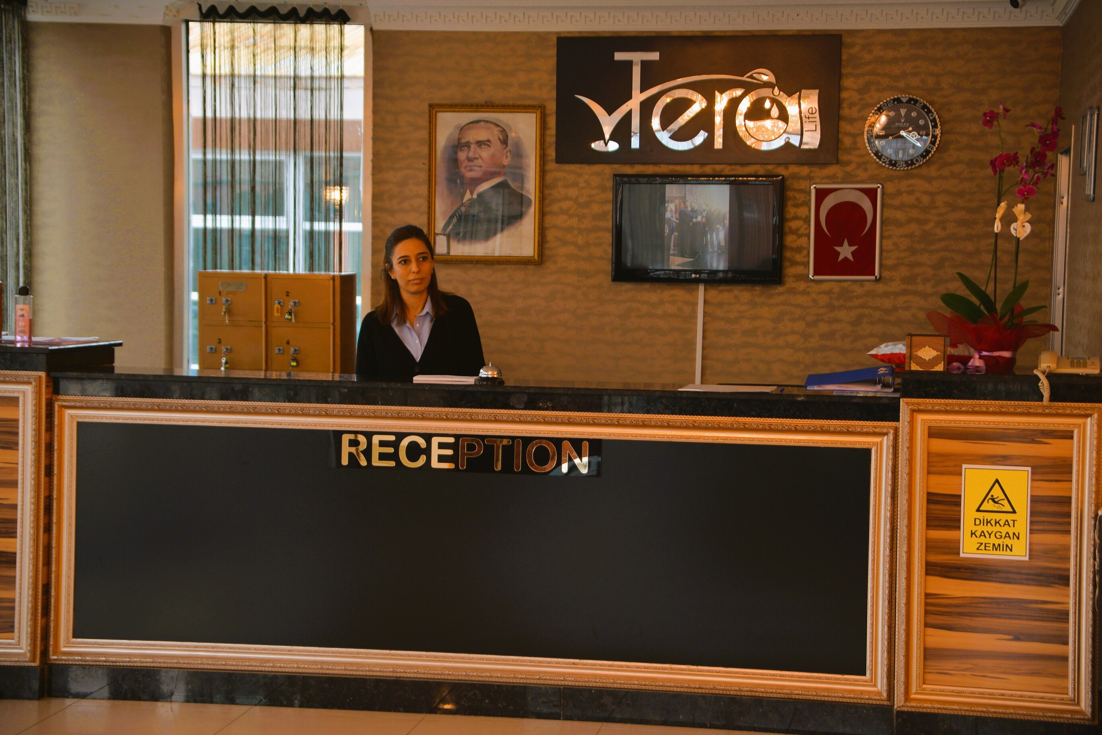 Een receptiebalie met een bordje 'RECEPTION', een lijst met een portret, een klok en een televisie aan de muur.