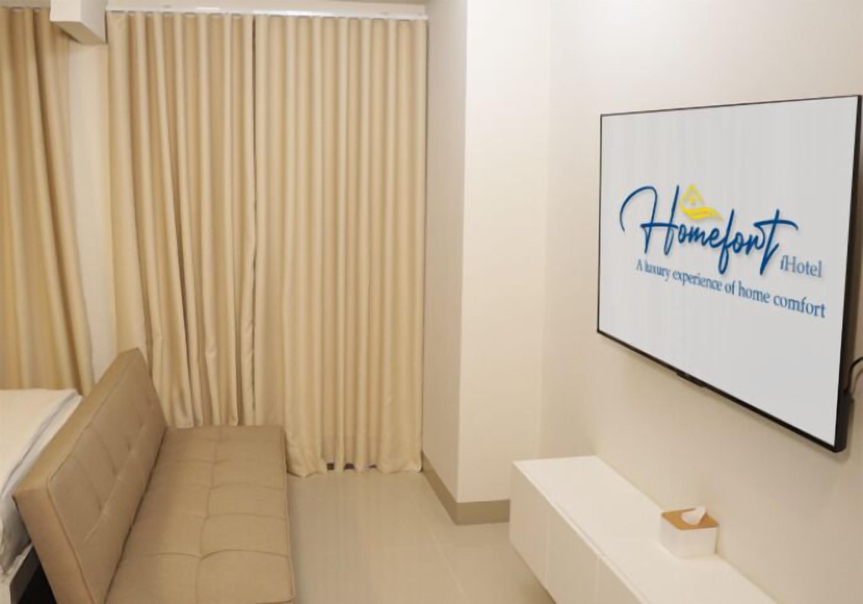 Ett hotellrum med en beige soffa, en platt-TV som är monterad på väggen och visar hotellets logotyp, och ett fönster med beige gardiner.